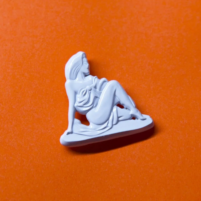 A small white figurine pin or charm shaped like a seated woman, placed on an orange background.