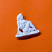A small white figurine pin or charm shaped like a seated woman, placed on an orange background.