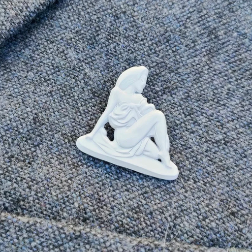 A small white figurine pin or charm shaped like a seated woman, placed on textured gray fabric.