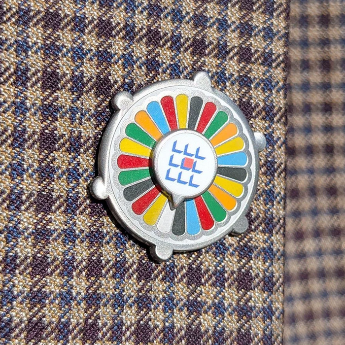A round enamel lapel pin with a silver-tone metal border, featuring a colorful radial pattern of red, yellow, green, blue, gray, and black segments around a central white circle with blue and red geometric symbols. The pin is displayed on a brown and navy plaid wool tie.