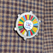 A round enamel lapel pin with a silver-tone metal border, featuring a colorful radial pattern of red, yellow, green, blue, gray, and black segments around a central white circle with blue and red geometric symbols. The pin is displayed on a brown and navy plaid wool tie.