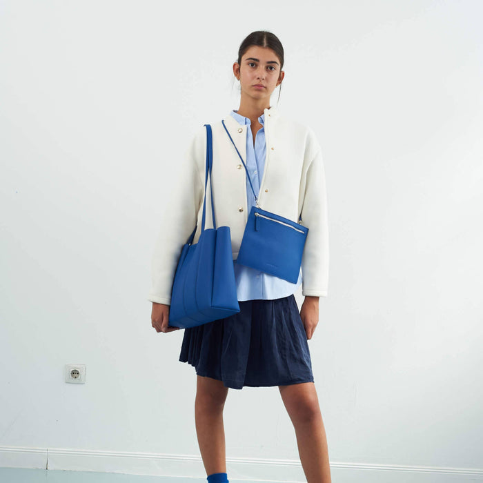 Person wearing a white blazer, blue skirt, and carrying two blue bags against a white background