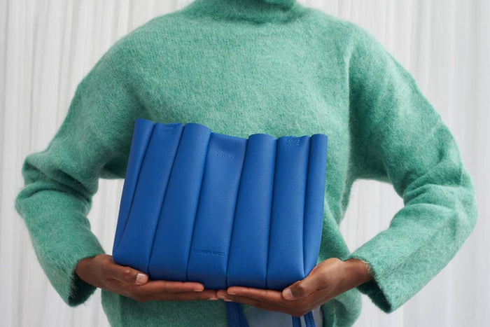 Person wearing a green sweater holding a blue pleated handbag against a white background