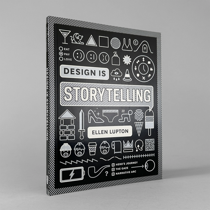 The book 'Design Is Storytelling' by Ellen Lupton, shown standing upright at a slight angle against a neutral gray background. The cover is black with white line-art illustrations and text.
