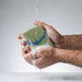 Hand holding a green sponge with soap suds under running water.