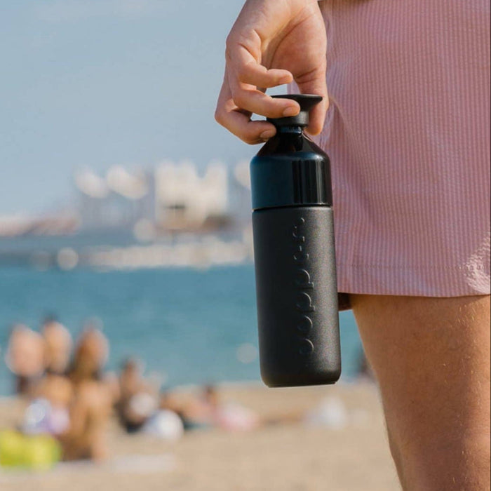 Person holding a black water bottle on a beach with people and a ship in the background