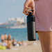 Person holding a black water bottle on a beach with people and a ship in the background