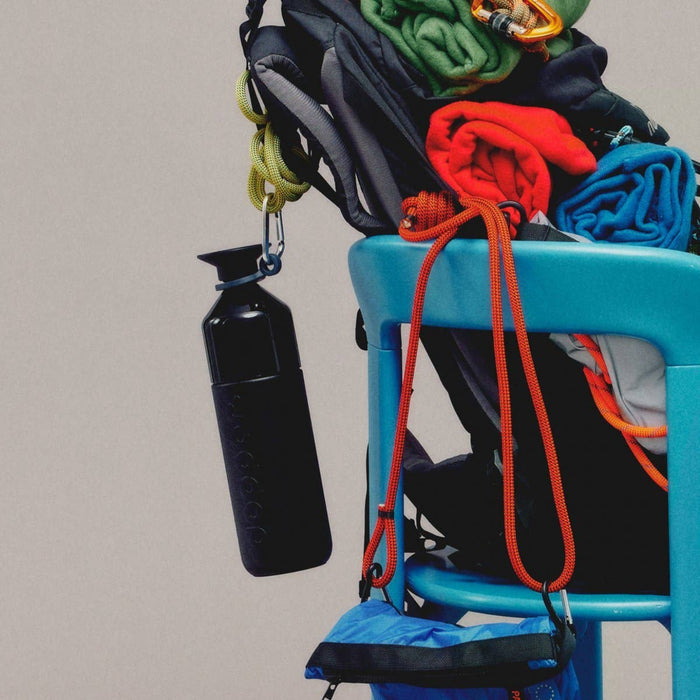 A black insulated water bottle with a carabiner clip hangs from a bright blue chair next to a pile of colorful rolled climbing or outdoor gear, including green, red, and blue fabric.