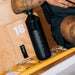 Person balancing an arm on the top of a black dopper water bottle with a wooden background.