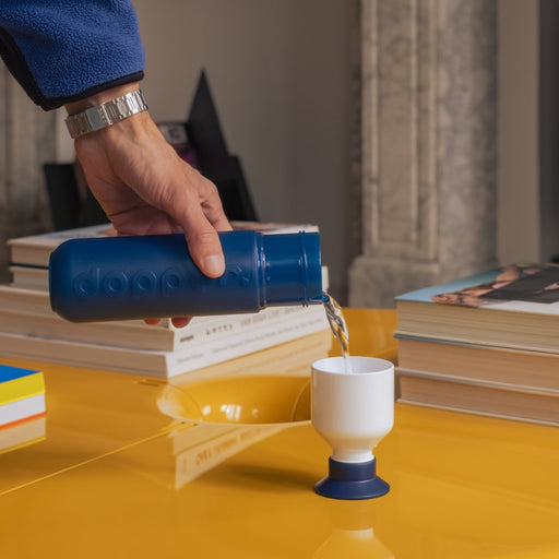 Hand pouring liquid from a blue container into a white cup on a yellow surface with books in the background.