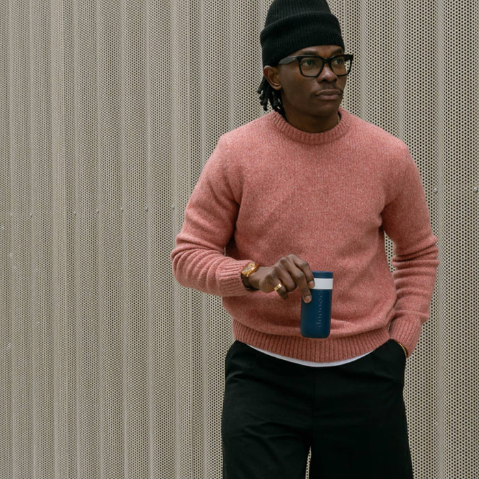 Person wearing a pink sweater and black pants, holding a blue cup against a textured wall.