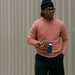Person wearing a pink sweater and black pants, holding a blue cup against a textured wall.