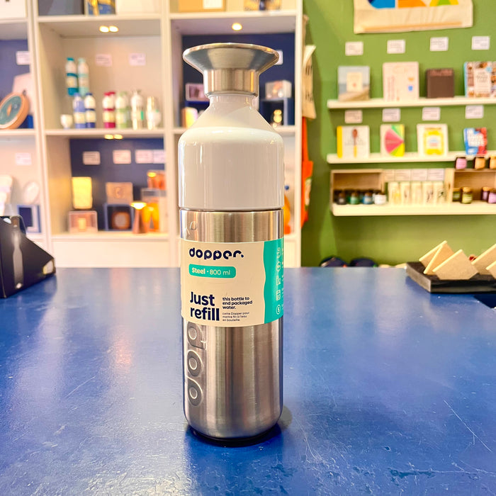 Silver and white reusable water bottle with 'Dopper' branding on a blue surface, with shelves in the background.