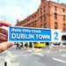 A hand holds a blue and white street sign reading 'Baile Atha Cliath DUBLIN TOWN 2' in front of a blurred brick building and city street background.