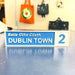 A rectangular street-style sign reading 'Baile Atha Cliath DUBLIN TOWN 2' in white text on a blue panel, placed on a reflective blue surface with a blurred indoor background and hanging bags behind it.