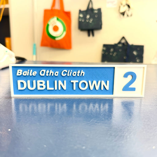 A rectangular street-style sign reading 'Baile Atha Cliath DUBLIN TOWN 2' in white text on a blue panel, placed on a reflective blue surface with a blurred indoor background and hanging bags behind it.