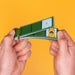 Two hands holding a green film strip camera that projects illustrated movie scenes, set against a solid yellow background.