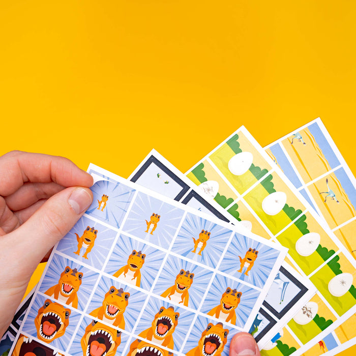 A hand holds a fanned-out set of colorful printed cards or stickers featuring cartoon orange dinosaur characters against a bright yellow background.