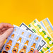 A hand holds a fanned-out set of colorful printed cards or stickers featuring cartoon orange dinosaur characters against a bright yellow background.