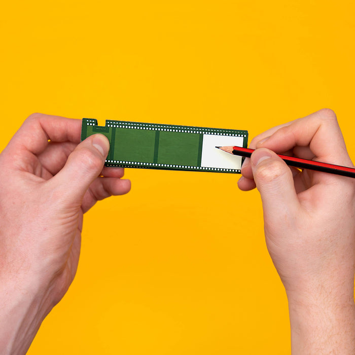 Two hands holding a green rectangular object against a bright yellow background, with a pencil being drawn on a white section of the object.