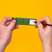 Two hands holding a green rectangular object against a bright yellow background, with a pencil being drawn on a white section of the object.
