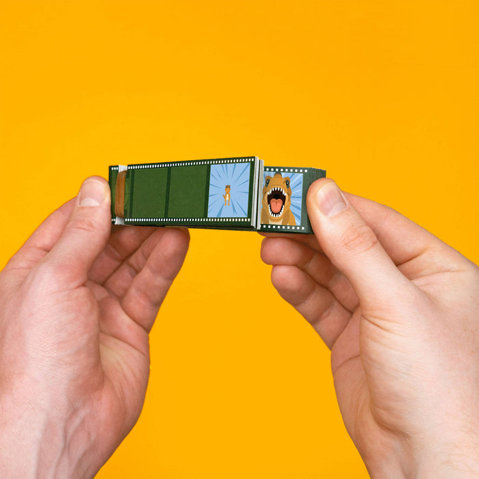 A miniature film strip, held between two hands against a solid bright yellow background. The flash drive features printed panels depicting a green film reel, a blue sky with a small figure, and a cartoon screaming face.