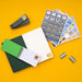 A flat-lay arrangement of sticker sheets, a small spiral notebook with a green cover, and several small rectangular sticker packs on a solid yellow background.