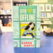 The book 'Effie Is Offline' by Karen Harte, featuring a light teal cover with comic-style illustrations and a central illustration of a girl in a plaid shirt and blue pants, standing on a blue table with a blurred bookstore background.