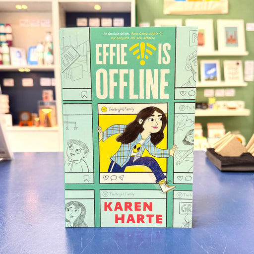 The book 'Effie Is Offline' by Karen Harte, featuring a light teal cover with comic-style illustrations and a central illustration of a girl in a plaid shirt and blue pants, standing on a blue table with a blurred bookstore background.