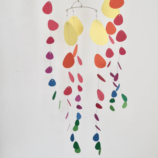 A hanging mobile with thin metal arches and dangling paper shapes in yellow, orange, pink, purple, blue, and green on a plain light background.