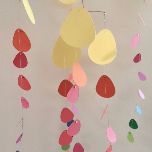 A hanging mobile with irregular pastel paper shapes in yellow, pink, red, blue, green, and purple, suspended from thin metal wires against a plain light gray wall.