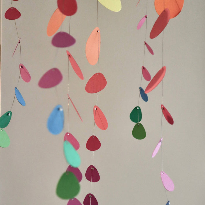 Colorful hanging paper leaf mobiles in various shapes and sizes, including green, pink, blue, orange, and yellow, suspended against a plain light gray wall.