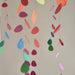 Colorful hanging paper leaf mobiles in various shapes and sizes, including green, pink, blue, orange, and yellow, suspended against a plain light gray wall.