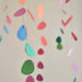 Colorful paper guitar-pick style hanging ornaments in pink, orange, yellow, blue, green, and purple suspended on strings against a plain light background.