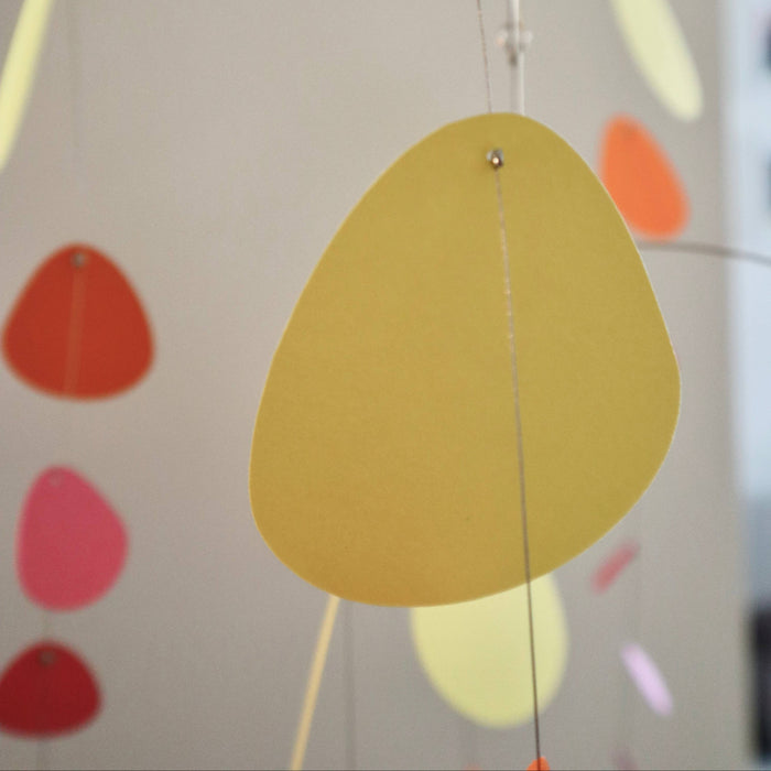 A modern mobile with a large yellow paper teardrop shape hanging from a thin metal frame, with other colorful paper shapes in orange, pink, and pale yellow blurred in the background.