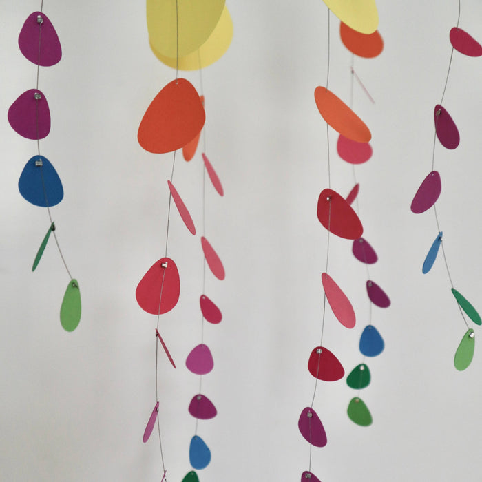 Colorful hanging paper garlands made of overlapping teardrop-shaped cutouts in yellow, orange, red, pink, blue, green, and purple, suspended against a plain light background.