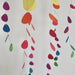 Colorful hanging paper garlands made of overlapping teardrop-shaped cutouts in yellow, orange, red, pink, blue, green, and purple, suspended against a plain light background.