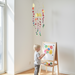 A young child stands at a small wooden easel painting on a colorful canvas in a bright, sunlit room with light wood floors and white walls. A multicolor hanging mobile hangs from the ceiling, and paint cups sit on the easel shelf.