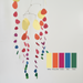 A colorful mobile wall hanging with curved metal wires and dangling paper-like shapes in yellow, orange-red, magenta, pink, blue, and green, shown against a plain light background with a color reference strip labeled Yellow, Orange-Red, Fuchsia, Blue, and Green.