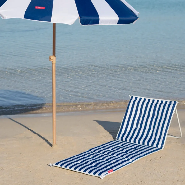 A blue and white striped beach umbrella and matching striped lounge chair on a sandy beach with the ocean in the background.