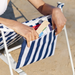 A close-up of a person putting a book into a blue and white striped zippered pouch that hangs on the back of a beach chair against a sand background.