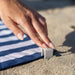 Hand placing a small white clip into sand next to a blue and white striped towel.