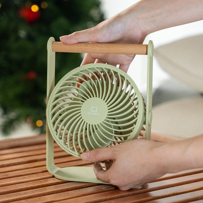 Person holding a small green fan on a wooden surface with a blurred Christmas tree in the background