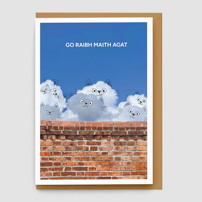 A greeting card with a brown envelope, featuring a blue sky, cartoon gray cats peeking over a red brick wall, and the text 'GO RAIBH MAITH AGAT' in white at the top.