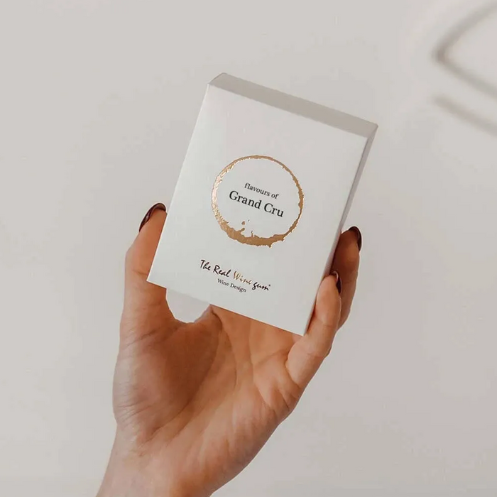 A hand holds a small white rectangular box with a gold circular logo reading 'Essence of Grand Cru' and smaller text 'The Real Wine Bar' on a light neutral background.