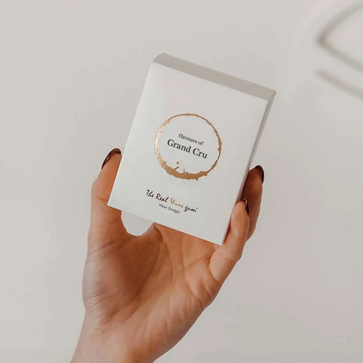 A hand holds a small white rectangular box with a gold circular logo reading 'Essence of Grand Cru' and smaller text 'The Real Wine Bar' on a light neutral background.