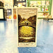 Decorative bookmark with a cut-out design of a bridge over water against a shop background.