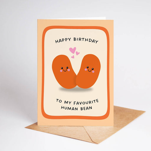 A birthday greeting card with a peach border and illustration of two smiling orange beans facing each other with small pink hearts. The text reads 'HAPPY BIRTHDAY TO MY FAVOURITE HUMAN BEAN'.