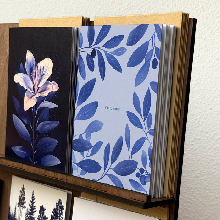 A stack of greeting cards displayed in a wooden card holder against a textured white wall. The front cards feature blue botanical leaf patterns, one in light blue with the text 'i'm so sorry', and another dark blue card with a large pale pink flower on a black background.