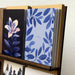 A stack of greeting cards displayed in a wooden card holder against a textured white wall. The front cards feature blue botanical leaf patterns, one in light blue with the text 'i'm so sorry', and another dark blue card with a large pale pink flower on a black background.
